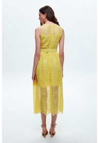 Gelbes Spitzenkleid mit tailliertem Schnitt und ausgestellt geschnittenem Saum. Aufwendige Blumenmuster überall, mit einem transparenten Rücken und einem Gürtel-Detail.