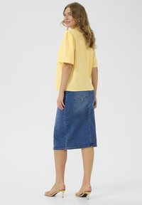 Haut jaune à manches courtes associé à une jupe midi en denim, de forme droite et avec des coutures subtiles. Complété par des sandales à talons jaunes.