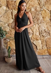 Schwarzes Maxikleid mit Neckholder, gerafftem Oberteil, fließender Silhouette und weichem Stoff; kombiniert mit hellbraunen Sandalen und erdigen Hintergrund.
