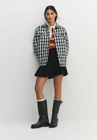 Sort-hvidternet oversized jakke, orange stribet top, sort plisseret nederdel, knælange sorte læderstøvler med hvid kant.