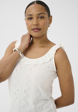 Femme aux cheveux foncés portant un haut blanc sans manches à volants, un bracelet argenté et des boucles d'oreilles en créole, regardant calmement la caméra.