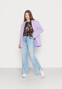 Blazer oversized de lavanda sobre uma blusa preta com estampa floral, combinada com jeans de perna larga em azul claro com fendas laterais e sapatilhas em tons pastel.