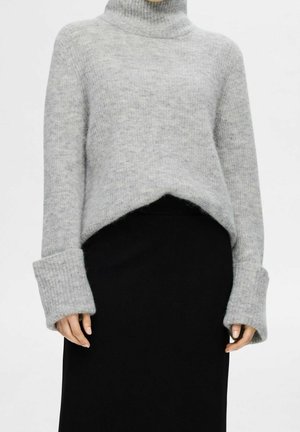 Person iført en lysegrå, blød rullekravetrøje med brede manchetter og et sort skørt mod en ensfarvet hvid baggrund.