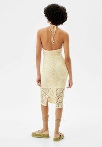Abito giallo in maglia con schiena scoperta, legacci al collo e design aderente. L'orlo presenta uno spacco e un motivo decorativo all'uncinetto. Abbinato a sandali verdi.
