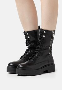 Botas de cuero negro con cordones y cremallera lateral, suela gruesa y accesorios de hardware plateado. Textura suave y altura hasta la mitad de la pantorrilla.