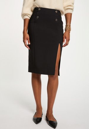 Gonna nera al ginocchio con spacco frontale e dettagli con bottoni, indossata con maglione crema, braccialetti dorati, anelli e ballerine nere appuntite.