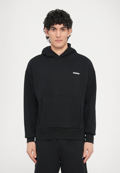 Melns sporta džemperis ar kapuci, priekšējo kabatu un ribveida aprocēm. Uz krūtīm maza balta logo, izgatavots no mīksta, strukturēta auduma.