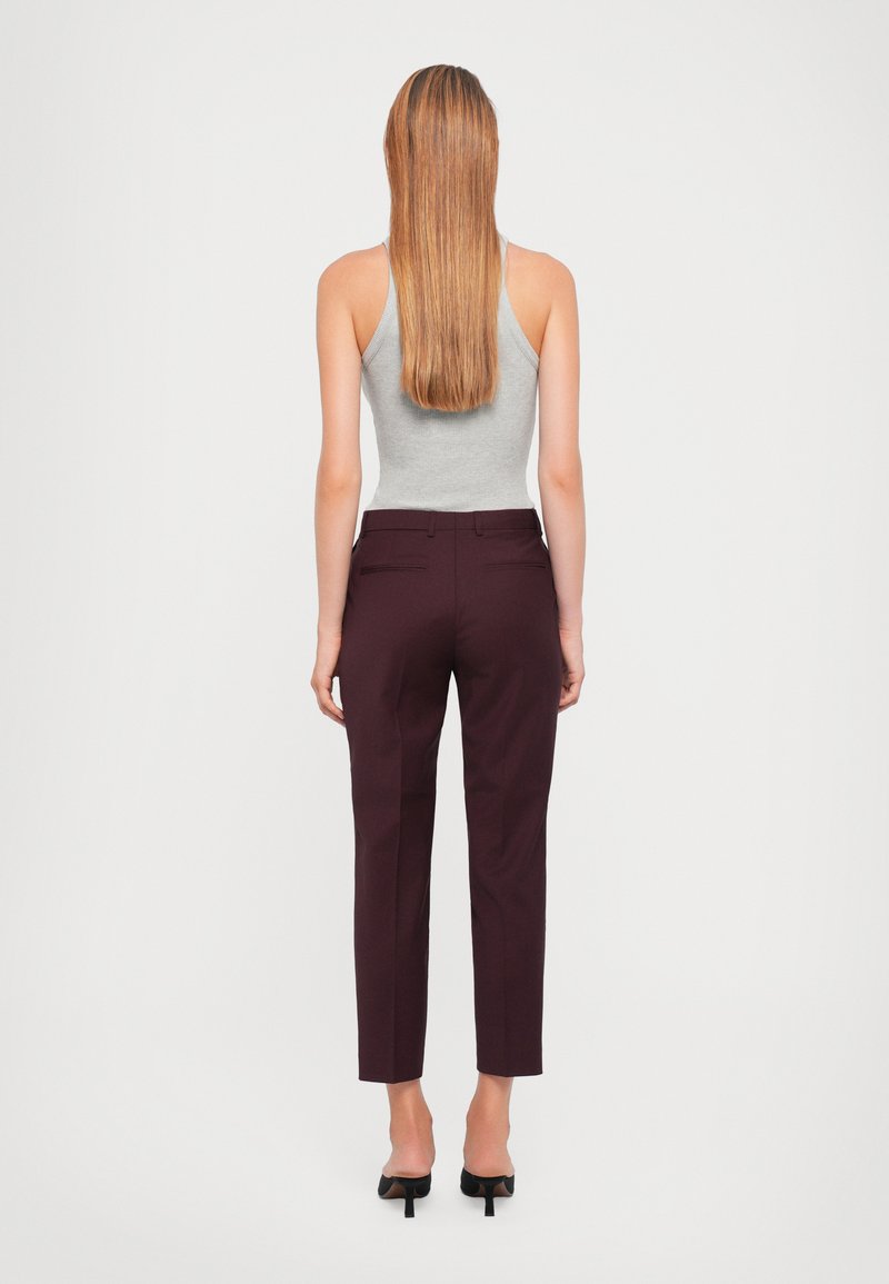 Grijze mouwloze top, bordeaux rode op maat gemaakte broek, achteraanzicht. De broek heeft achterzakken en taps toelopende pijpen. Het model draagt zwarte hoge hakken.