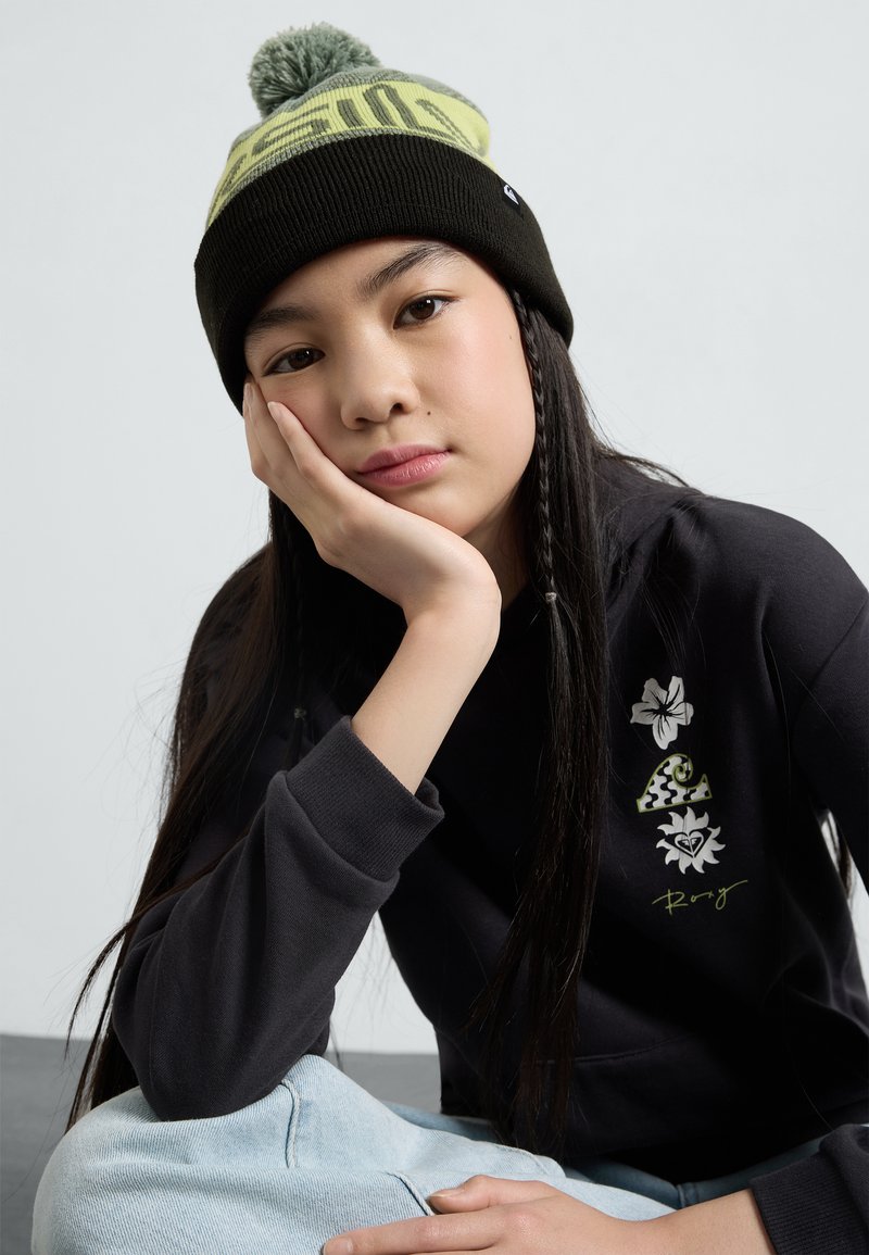 Haut à capuche noir avec broderie florale, jeans bleu clair et bonnet en tricot noir avec un pompon vert et un texte graphique.