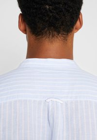 Chemise rayée bleu clair avec une texture douce, dotée d'un détail en boucle à l'arrière du col et d'une coupe décontractée.