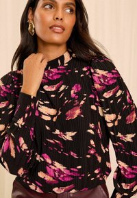 Schwarze Bluse mit langen Ärmeln, plissierter Textur und floralem Muster in Pink und Creme. Hoher Kragen und geknöpfte Ärmel.