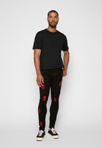 Melnas īsām piedurknēm T-krekls, pieguļošs siluets. Šauras melnas džinsu bikses ar sarkanu galvaskausu un krustotu kaulu izšuvumu. Ikdienas melnas sporta apavus ar krēmkrāsas zoli.