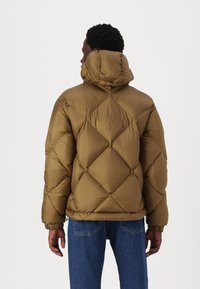 Giacca a vento marrone con un design a rombi, cappuccio e polsini elasticizzati. Il materiale appare liscio con una finitura leggermente lucida.
