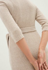 Abito beige a coste con maniche a tre quarti e una cintura abbinata in vita. Il tessuto ha un aspetto morbido e strutturato.