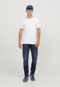 Camiseta blanca de manga corta, vaqueros azul oscuro y una gorra azul con escritura. Los zapatos son azules con acentos texturizados y una suela de goma marrón.