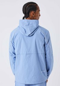 Hombre con cabello trenzado que lleva una chaqueta con capucha de color azul claro con detalles de ribete blanco, de pie de espaldas contra un fondo blanco.