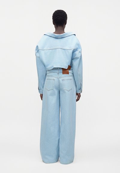 Personne aux cheveux courts portant une veste en jean courte bleu clair et un jean taille haute à jambes larges vue de dos sur un fond uni.