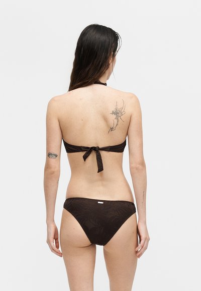 Γυναίκα με μακριά σκούρα μαλλιά που φοράει μαύρο μπικίνι, δείχνει την πλάτη της, με τατουάζ στο πάνω δεξί ώμο και αριστερό μπράτσο.