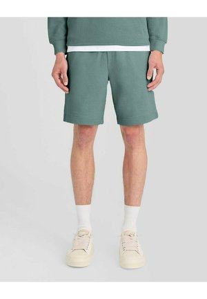 Man draagt groene casual shorts, witte sokken, beige sneakers en een bijpassende groene trui, staand tegen een effen achtergrond.