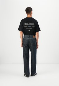 La camiseta negra presenta texto blanco en la parte posterior con un logo. Se lleva con jeans de mezclilla azul oscuro y zapatos negros con rayas blancas.