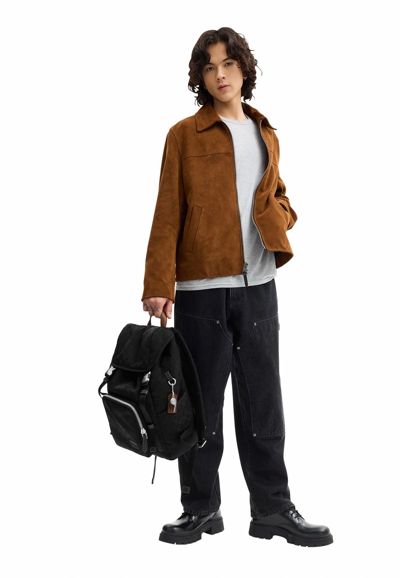 Young person with curly hair wearing a brown jacket, gray shirt, black pants, and boots holding a black backpack by the handle.