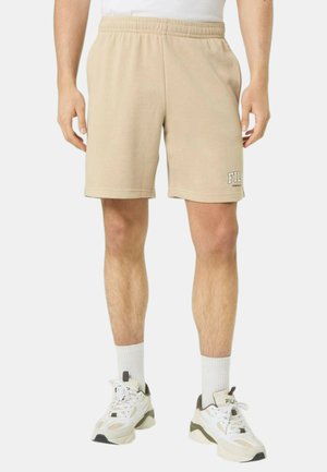 Uomo che indossa pantaloncini in pile beige FILA, calzini bianchi al polpaccio e sneaker bianche FILA, in piedi contro uno sfondo bianco uniforme.