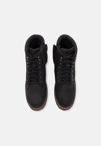 Botas de ante negro con cordones, cuellos acolchados, ojales de metal y suela de goma marrón en contraste. Diseñadas con una textura suave.