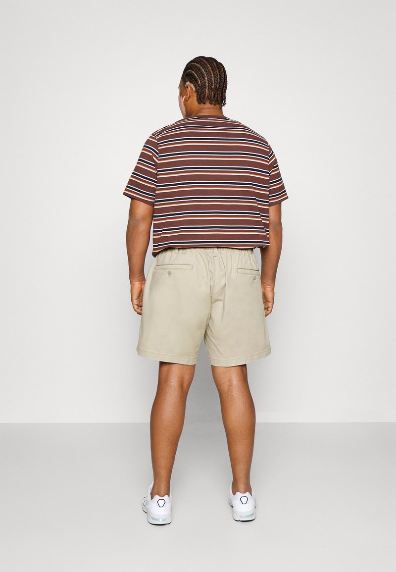 Mann mit geflochtenem Haar, der ein braun gestreiftes T-Shirt, beige Shorts und weiße Turnschuhe trägt und vor einer schlichten weißen Wand steht.