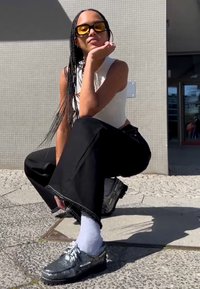 Schwarze weit geschnittene Hosen, weißes ärmelloses Top, silberne Schuhe, weiße Socken und gelb getönte Sonnenbrille. Texturierten Pflasterhintergrund.