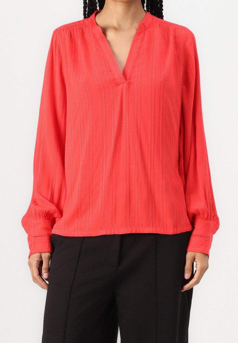 Femme portant un chemisier corail vif à manches longues, col en V avec des motifs verticaux subtils, associé à un pantalon noir.