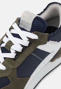 Αθλητικά παπούτσια που διαθέτουν μείγμα υλικών, συμπεριλαμβανομένου του ναυτικού υφάσματος, ελιάς σουέτ και λευκών δερμάτινων λεπτομερειών, με λευκές κορδόνες και καουτσούκ σόλα.