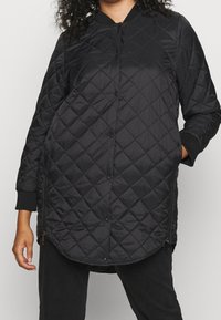 Vrouw in een lange zwarte gewatteerde jas met drukknoopjes, zijritsen, geribde manchetten en handen in de zakken.