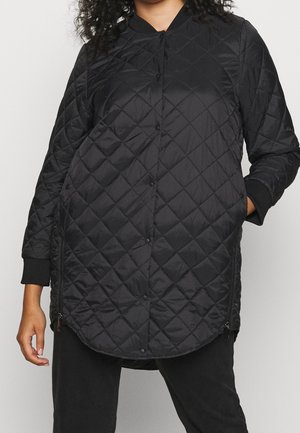 Kvinna som bär en lång svart vadderad jacka med tryckknappar, sidizipper, ribbade manschetter och händerna i fickorna.