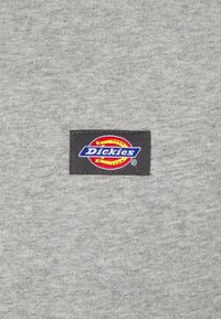 Un tessuto grigio con una consistenza morbida presenta un'etichetta nera cucita sopra, che mostra il logo "Dickies" in colori rosso, giallo e blu.