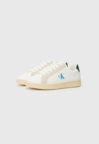 Baskets blanches avec des accents beiges et une languette de talon verte, arborant un logo bleu et des lacets plats, conçues avec une texture synthétique lisse.