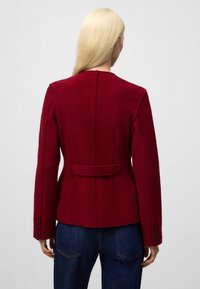 Czerwona wełniana kurtka z długimi rękawami, dopasowanym krojem, paskiem z tyłu i zapinanymi mankietami, w połączeniu z niebieskimi dżinsami.