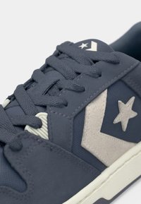 Sneaker featuring a dark blue suede upper, white star and chevron logo accents, rounded toe, and textured laces. Rubber sole visible.