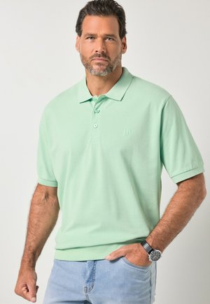 Homme barbu portant un polo vert clair et un jean bleu clair, une main dans la poche et une montre au poignet gauche.