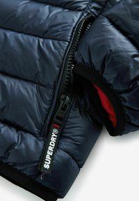 Chaqueta acolchada azul marino con un acabado brillante, que presenta una cremallera negra y un forro rojo, marcada con una etiqueta "SUPERDRY" en el tirador de la cremallera.