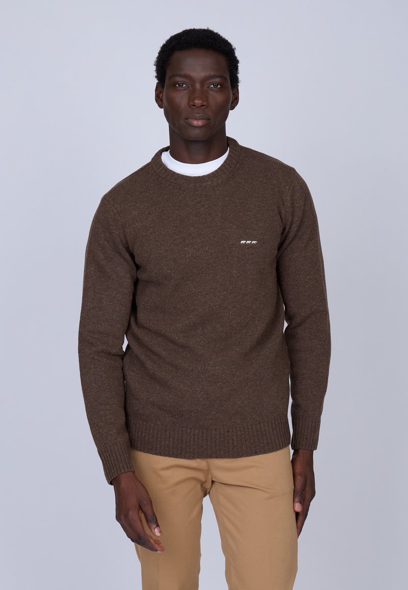 Pull en maille marron avec un col rond, tissu texturé, arborant un petit logo sur le côté gauche. Associé à un pantalon beige clair.