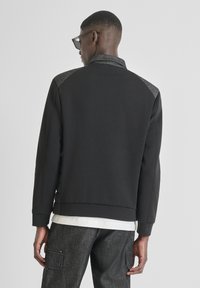 Felpa nera con colletto e pannelli delle spalle grigi a contrasto, texture liscia e polsini a coste. Indossata sopra una camicia bianca.