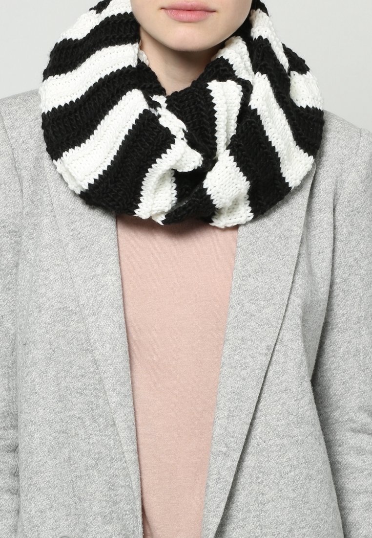 Personne portant une écharpe tricotée infinie rayée noire et blanche sur un haut rose clair et un manteau gris, visible du cou au milieu du torse.