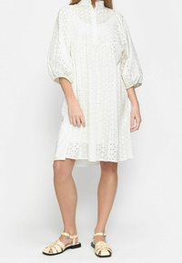 Vestido blanco de mangas abullonadas con patrones de ojetes, cuello alto y corte relajado. Llevado con sandalias beige de tiras. Textura de tela suave.