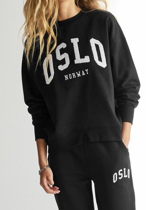 Kvinde iført sort sweatshirt og bukser med hvide bogstaver "OSLO", "NORWAY" tekst på sweatshirten, står med den ene hånd i lommen.