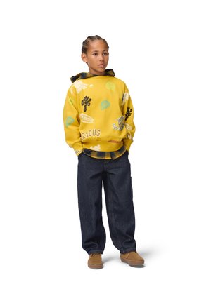 Enfant aux cheveux tressés portant un sweat à capuche jaune imprimé par-dessus une chemise à carreaux, un jean foncé et des chaussures marron, debout les mains dans les poches.