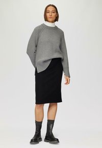 Vrouw die staat en een grijze trui over een witte coltrui draagt, een zwarte rok tot op de knie, donkere sokken en zwarte enkellaarsjes op een effen achtergrond.
