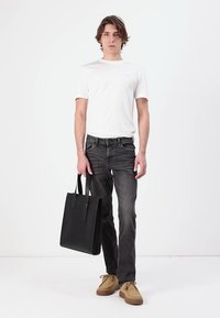 Sac fourre-tout en cuir noir avec deux poignées supérieures, texture lisse. Modèle portant un t-shirt blanc, un jean gris et des chaussures beiges. Design simple et moderne.