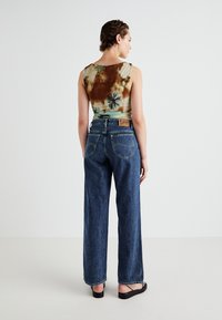 Jeans in denim scuro a gamba larga con cuciture gialle a contrasto e un'etichetta in pelle sul retro; modello che indossa una canotta tie-dye multicolore.