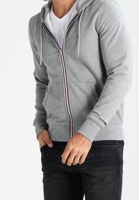 Grau zip-up Hoodie mit Kapuze, Fronttaschen und kontrastierendem Reißverschluss. Mit gerippten Bündchen und Saum. Getragen über einem weißen T-Shirt.