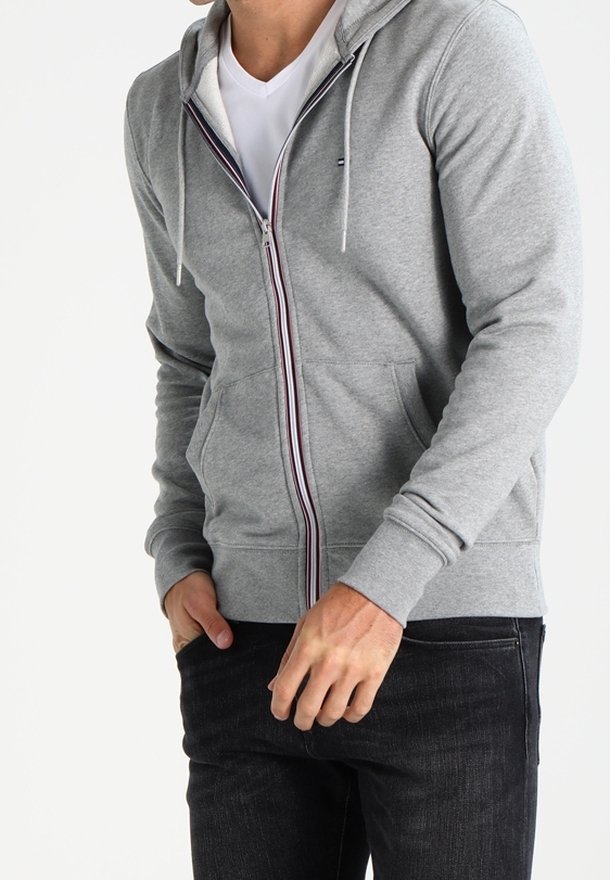 Grau zip-up Hoodie mit Kapuze, Fronttaschen und kontrastierendem Reißverschluss. Mit gerippten Bündchen und Saum. Getragen über einem weißen T-Shirt.